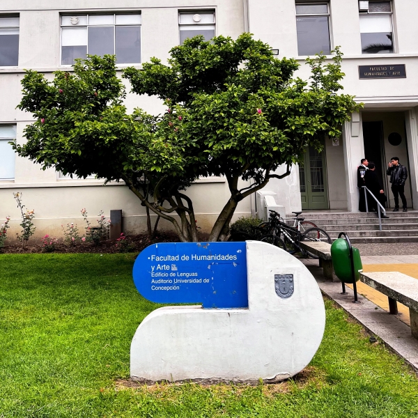 Two people stand in the doorway to an academic building at Universidad de Concepcion. A sign and a tree are outside.